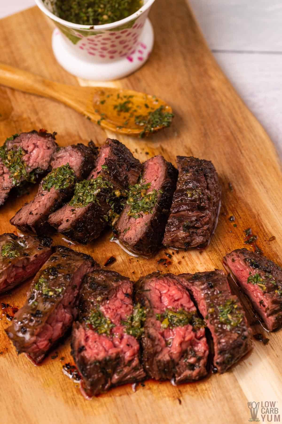 chimichurri flank steak sliced on cutting board with sauce container.