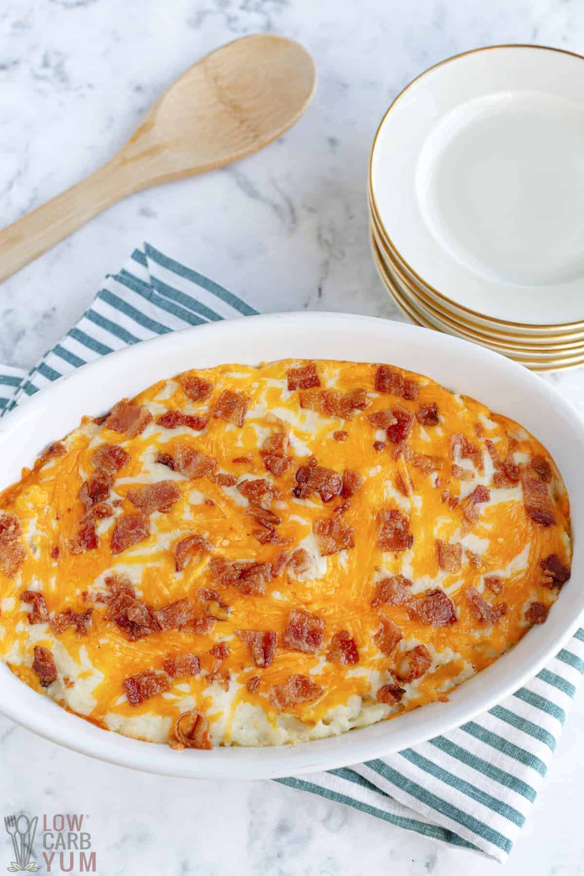 baked loaded cauliflower casserole in baking dish with wooden spoon and plates.