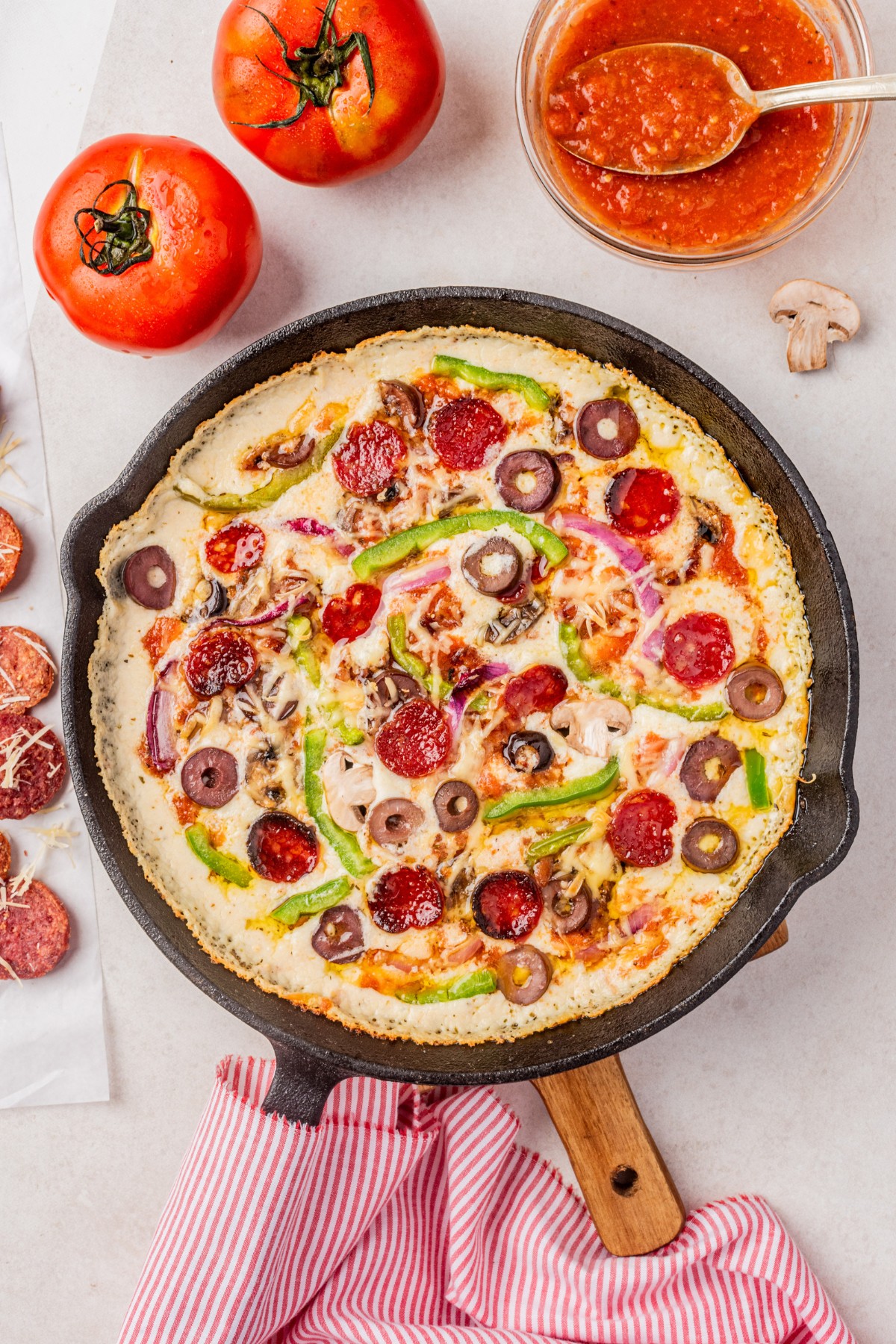 بالای سر پیتزا کتو بعد از پخت در یک تابه چدنی با حوله آشپزخانه دور دسته و سایر مواد روی میز قرار دهید.