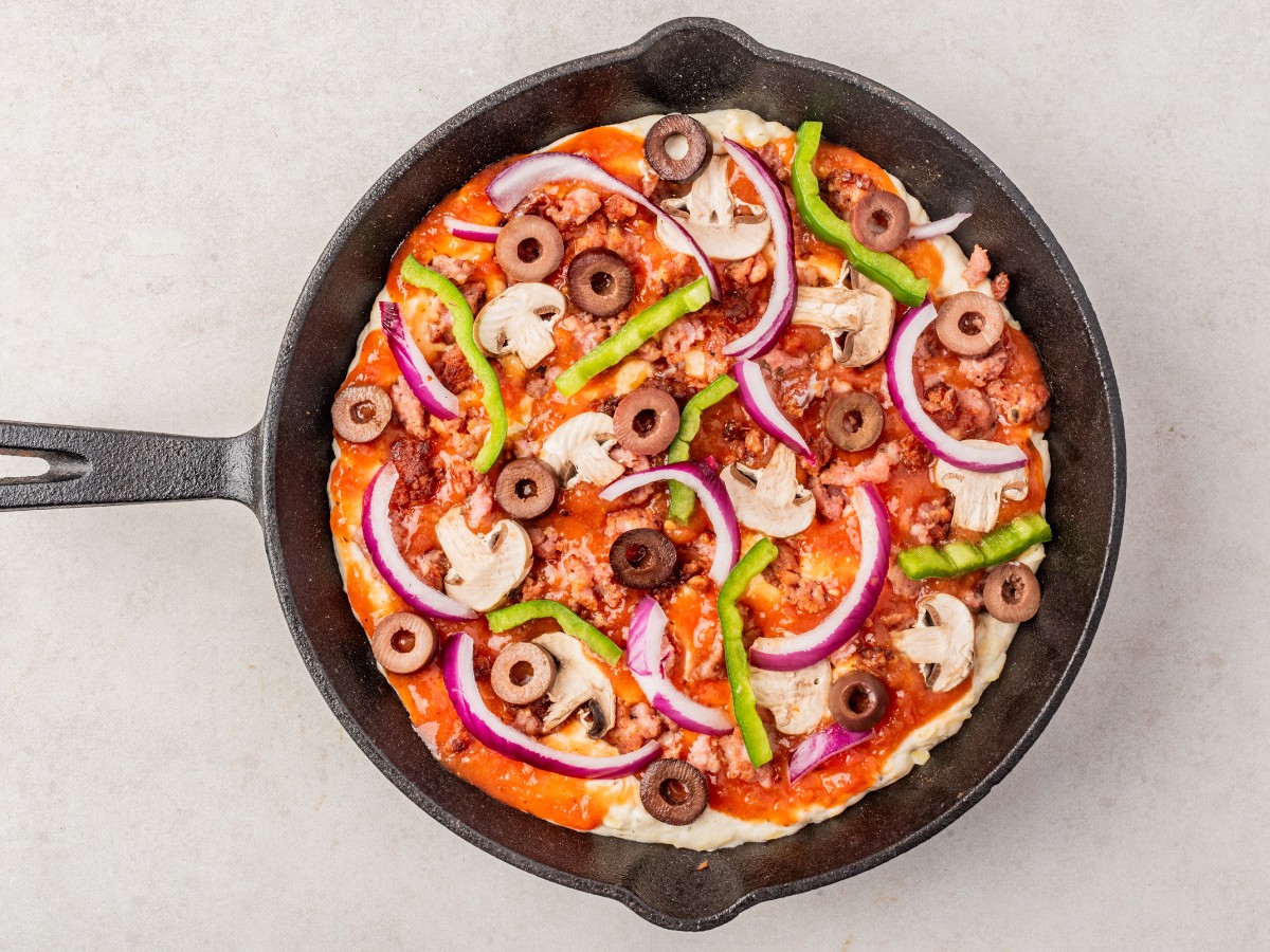 overhead of unbaked keto pizza dip in a cast iron pan with sauce and veggies added.