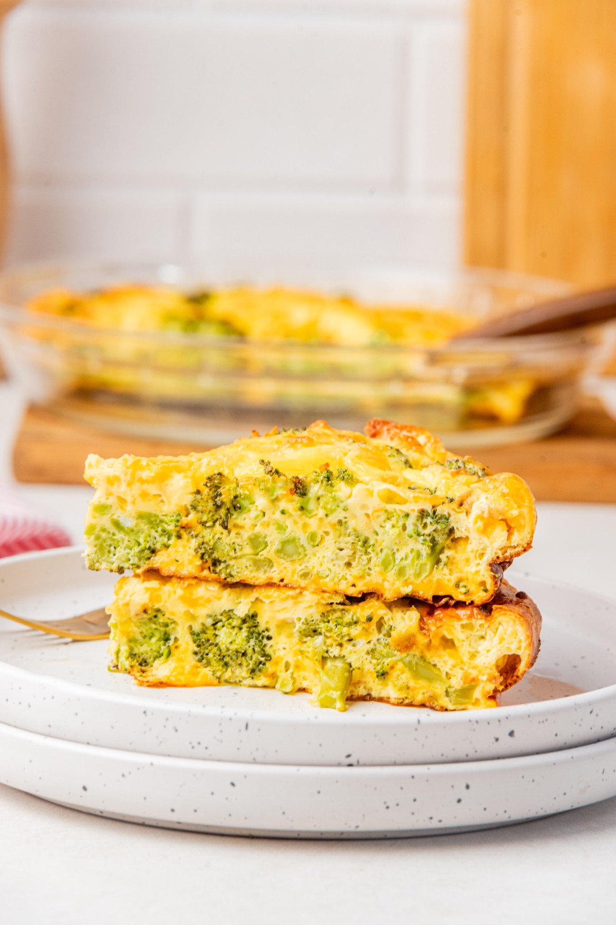 two slices of crustless broccoli cheddar quiche stacked on top of each other on two white plates with a fork.