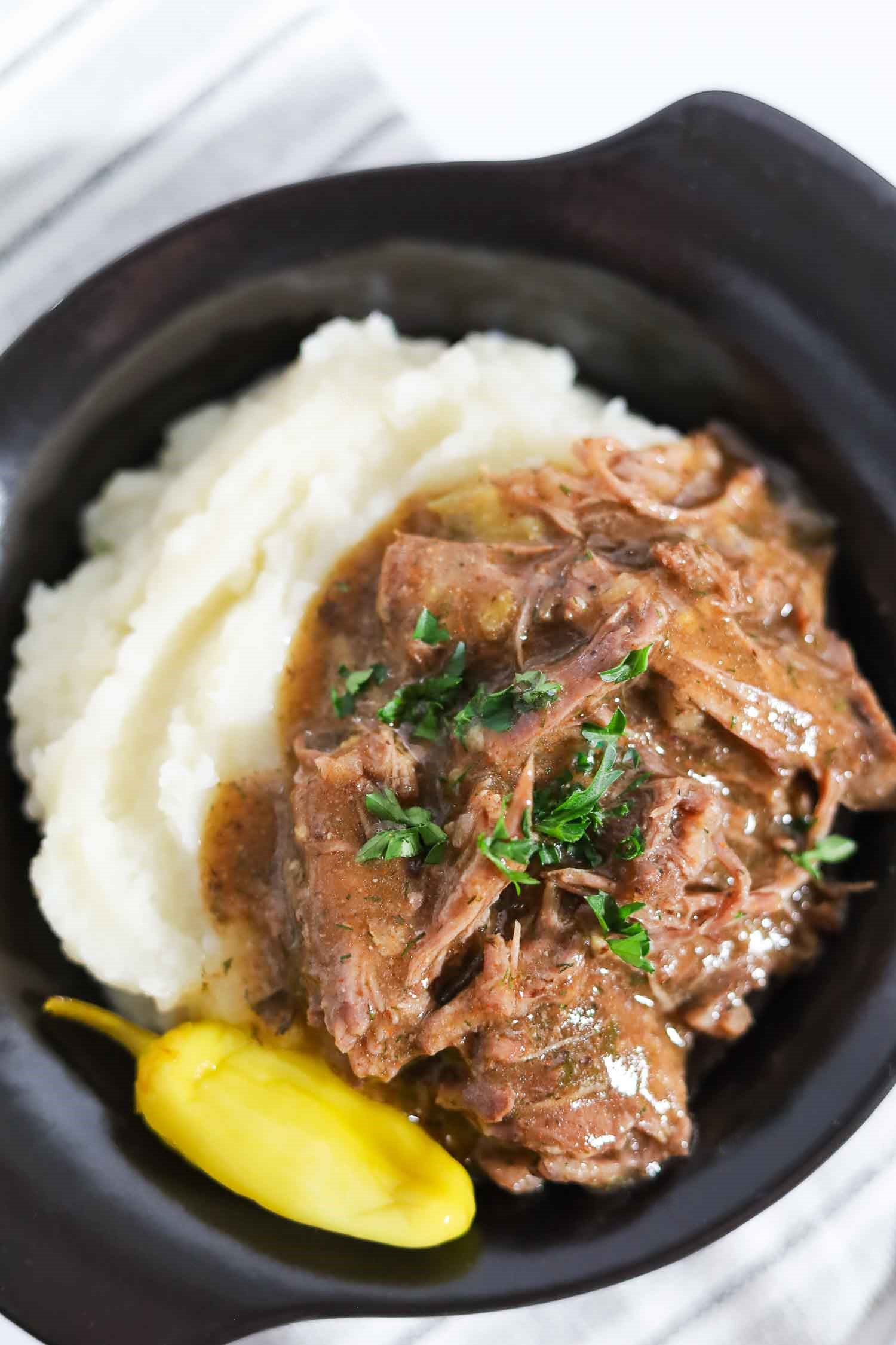 close-up shot of serving of slow cooker Mississippi pot roast in a black bowl with mashed potatoes and a pepperoncini pepper.