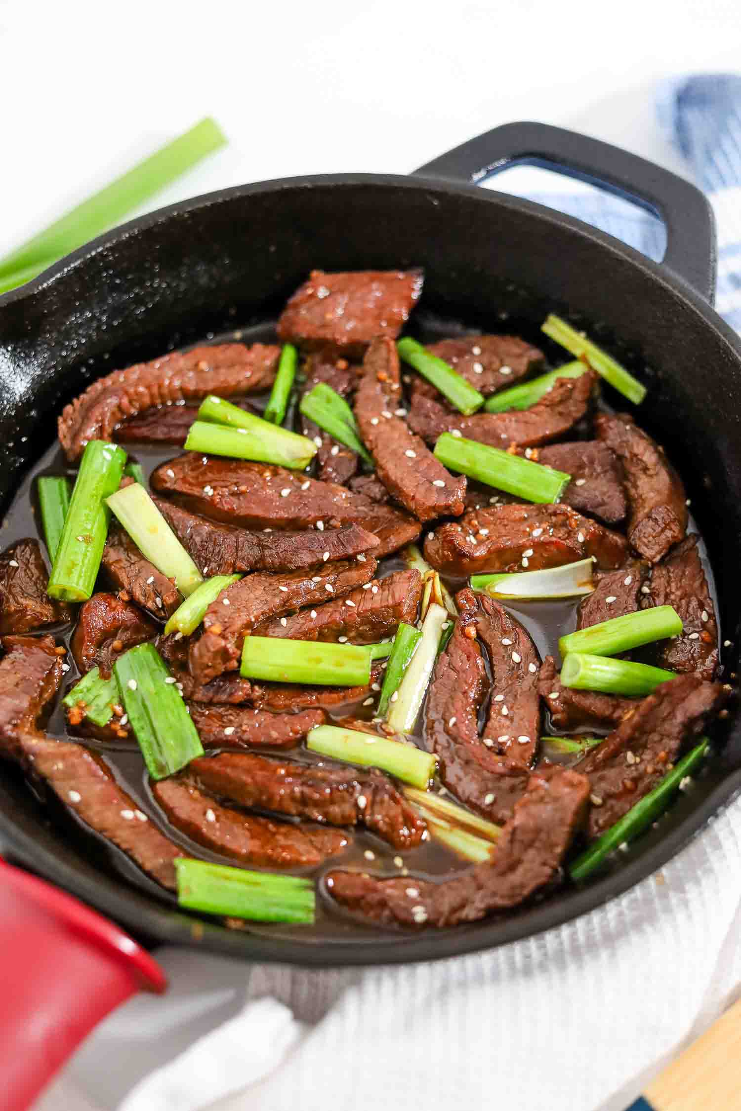 cooked keto mongolian beef in a skillet garnished with green onions and sesame seeds.